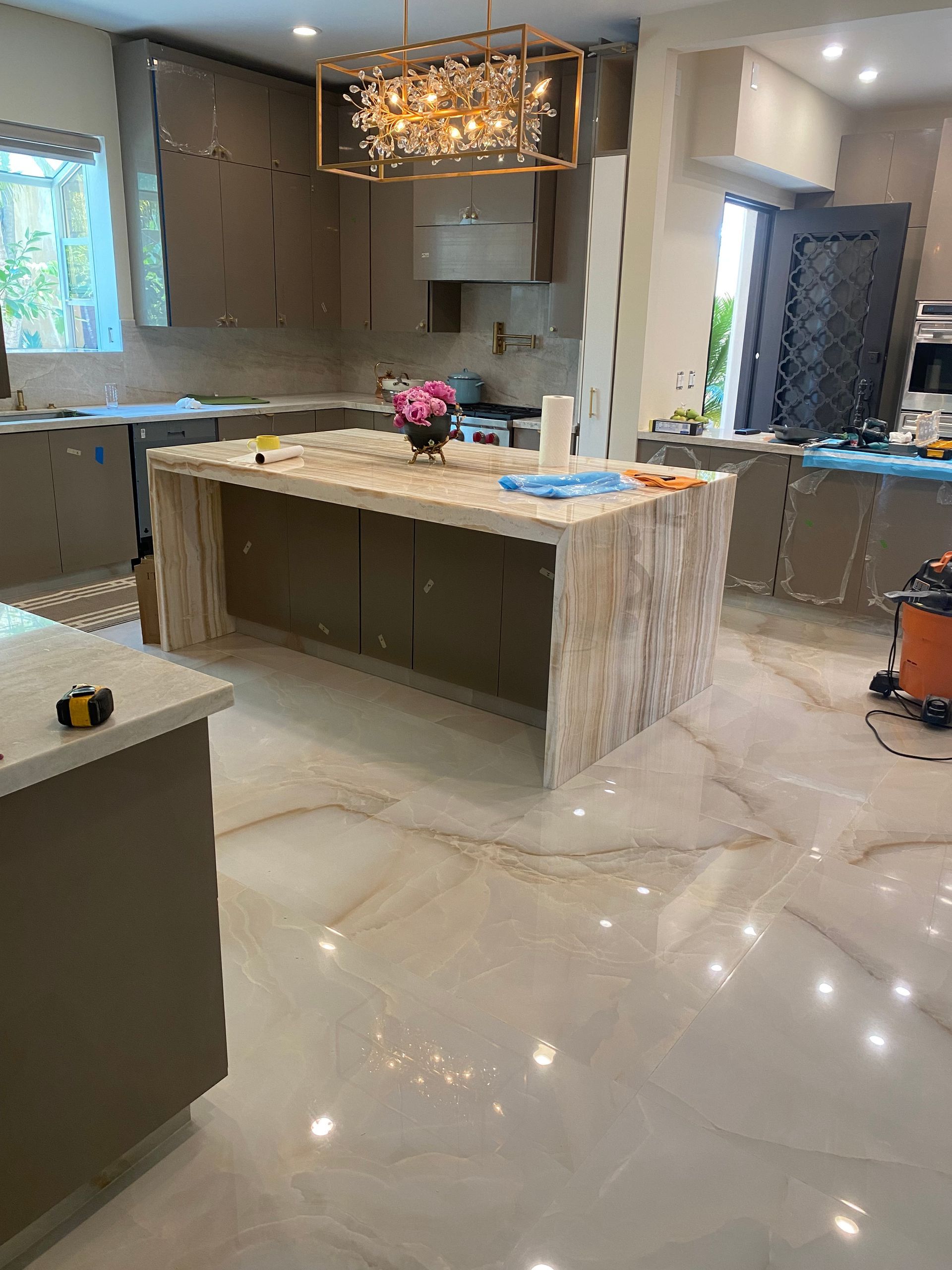 Modern kitchen with marble island, glossy floor, chandelier, and windowed counters; cleaning items on the island
