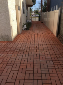 Narrow brick-paved walkway beside a beige building and fence, with potted plants along the left side.