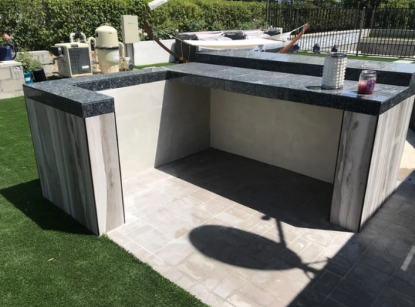 Outdoor built-in bar with black stone countertop, light stone base, and stools on a patio lawn area