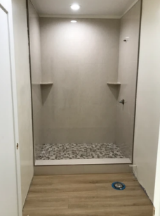 Empty shower stall with gray walls, built-in shelves, pebble floor, and wood-look flooring outside