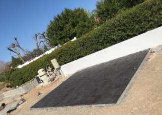Tilted view of a large black solar panel on a dirt lot beside a white wall and green trees
