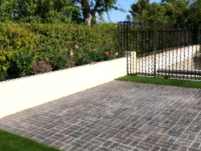Paved patio beside a white wall and black gate, with hedges and trees in the background.
