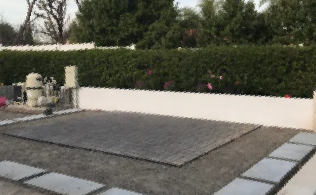 Stone patio with a white retaining wall, dense green hedge, and outdoor fountain in a garden.