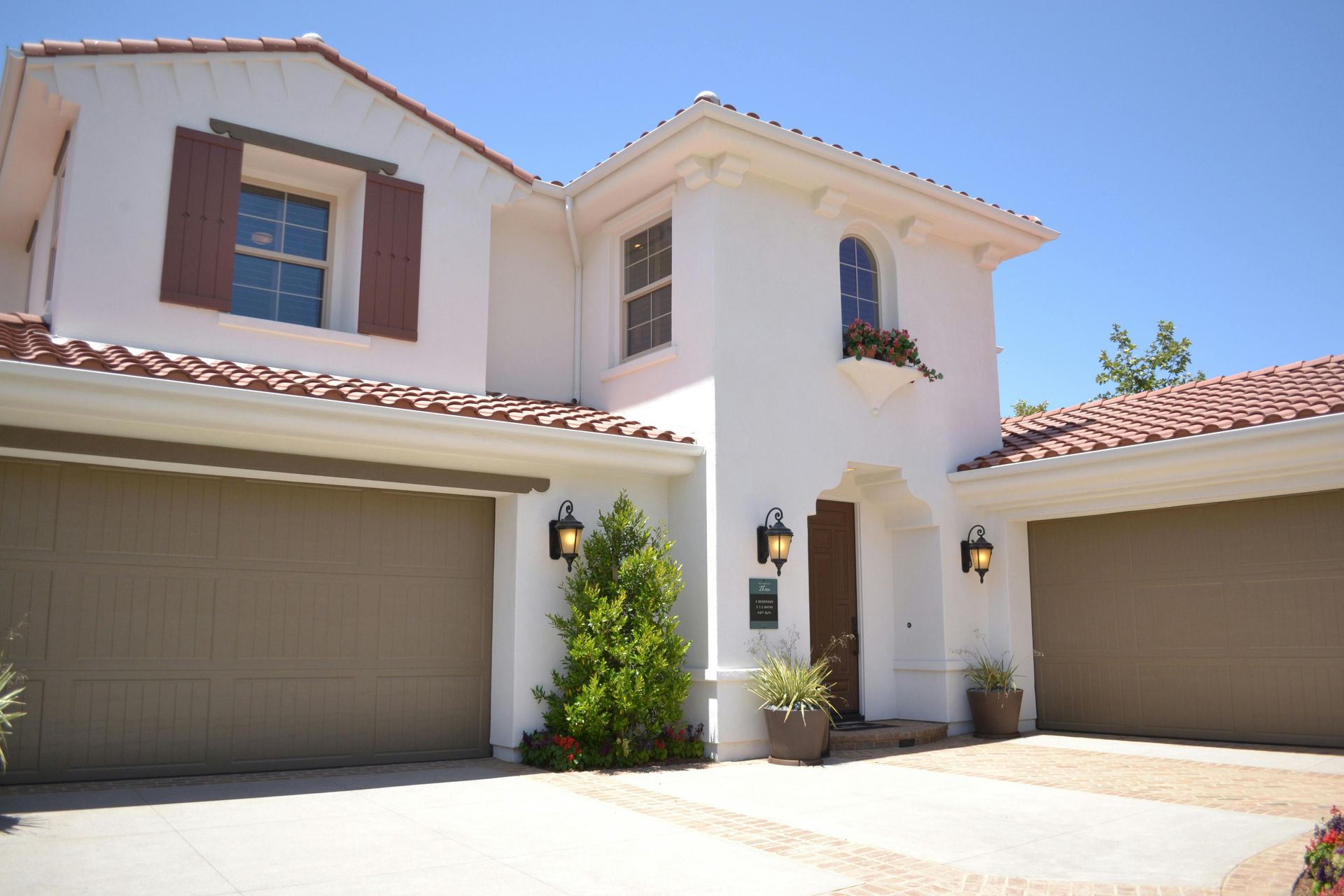 Front view of a beige stucco townhouse with red tile roofs and two garage doors under a bright blue sky