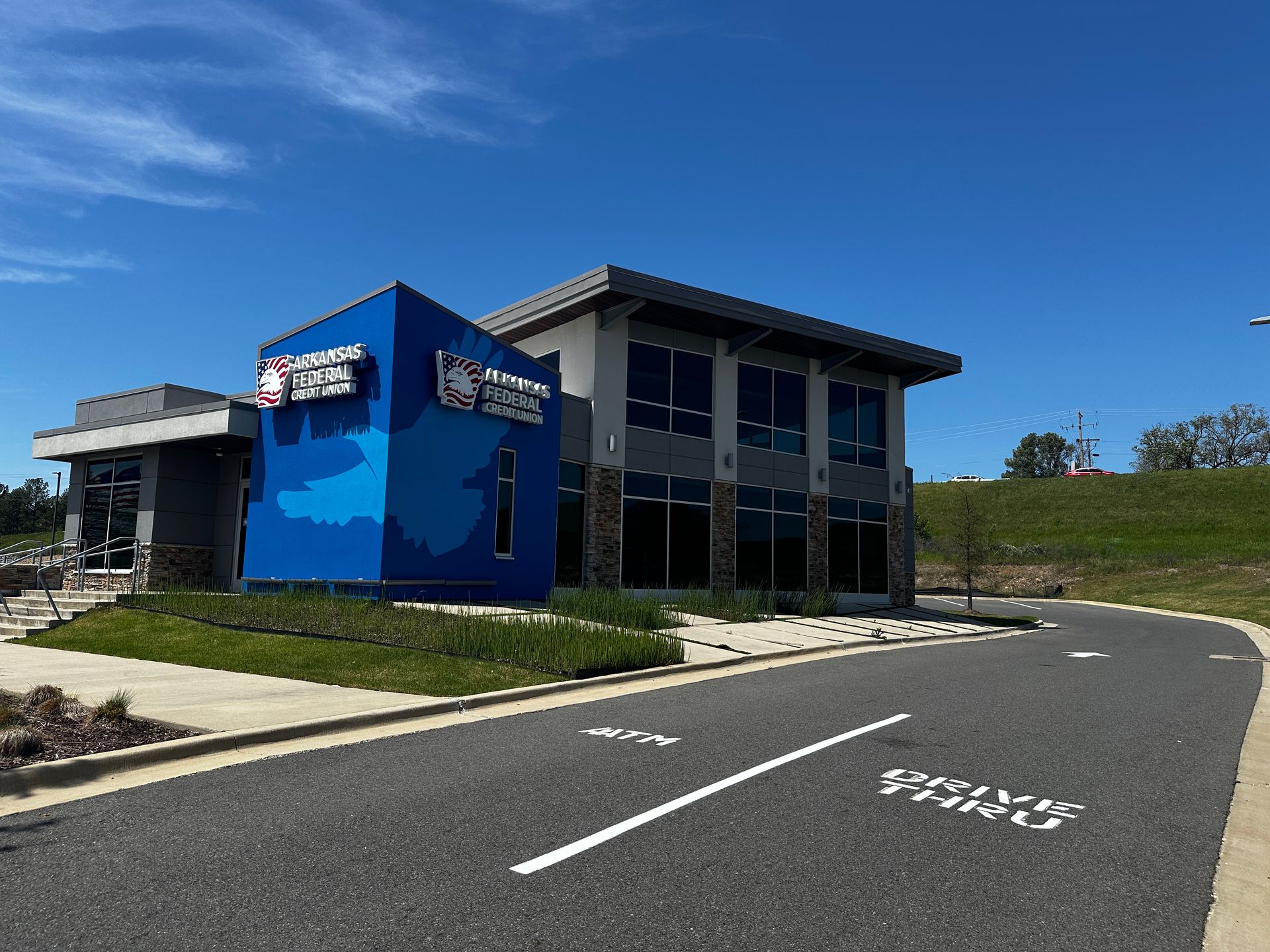 Two-story building with a blue facade, next to a parking lot and grassy hill under a blue sky.