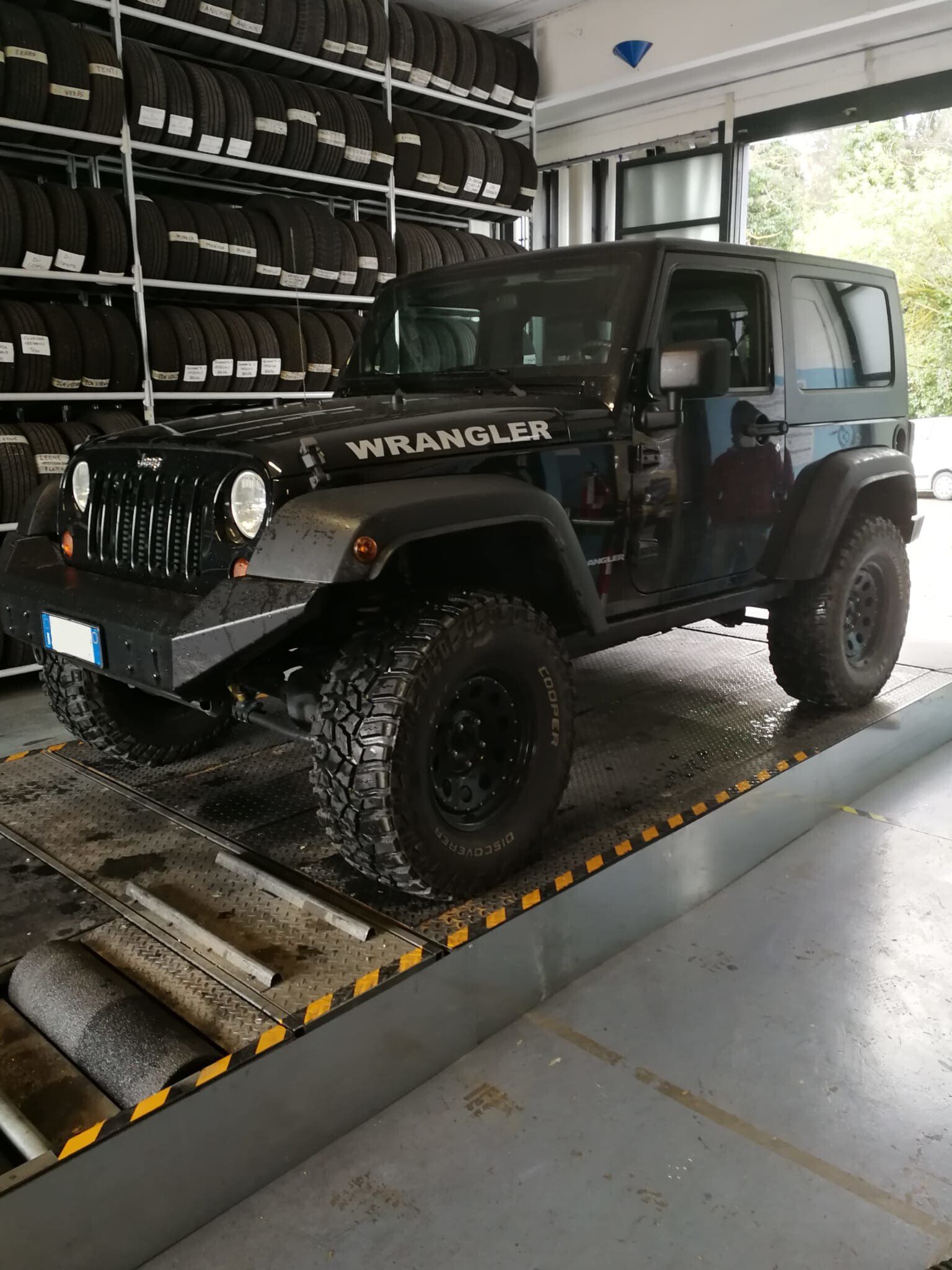 Una Jeep Wrangler nera su un ponte sollevatore in un garage, con pneumatici accatastati sugli scaffali.