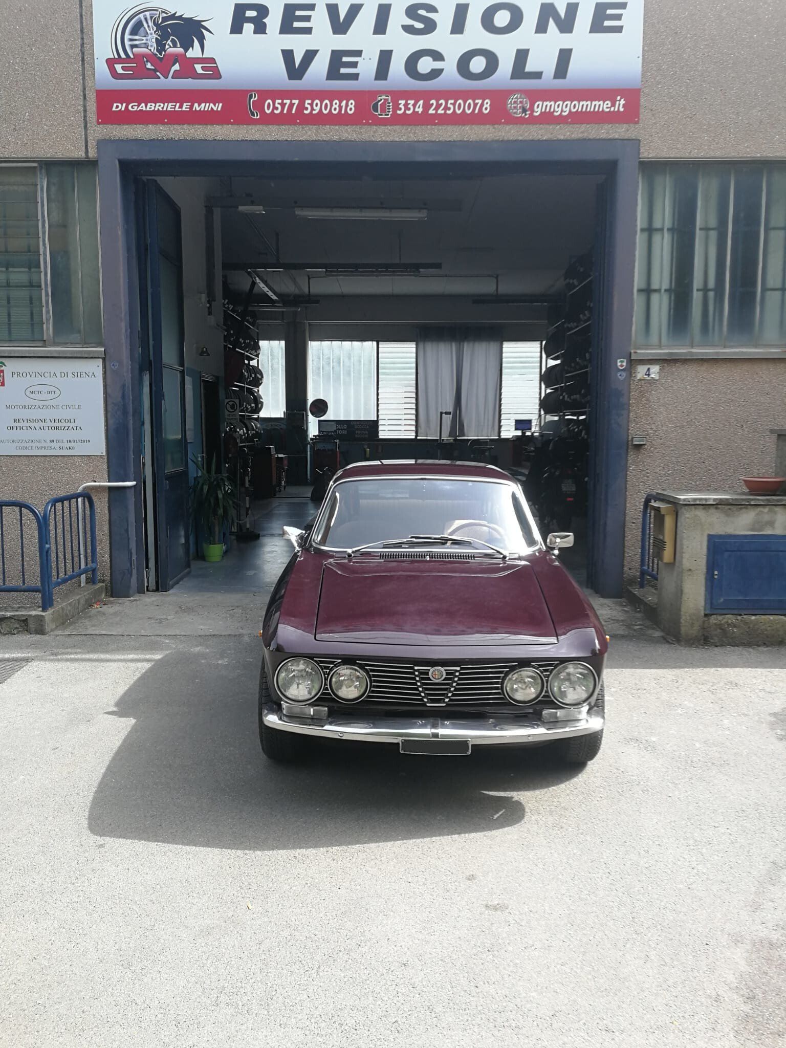 Un'auto d'epoca color Borgogna parcheggiata davanti a un'officina di revisione veicoli.