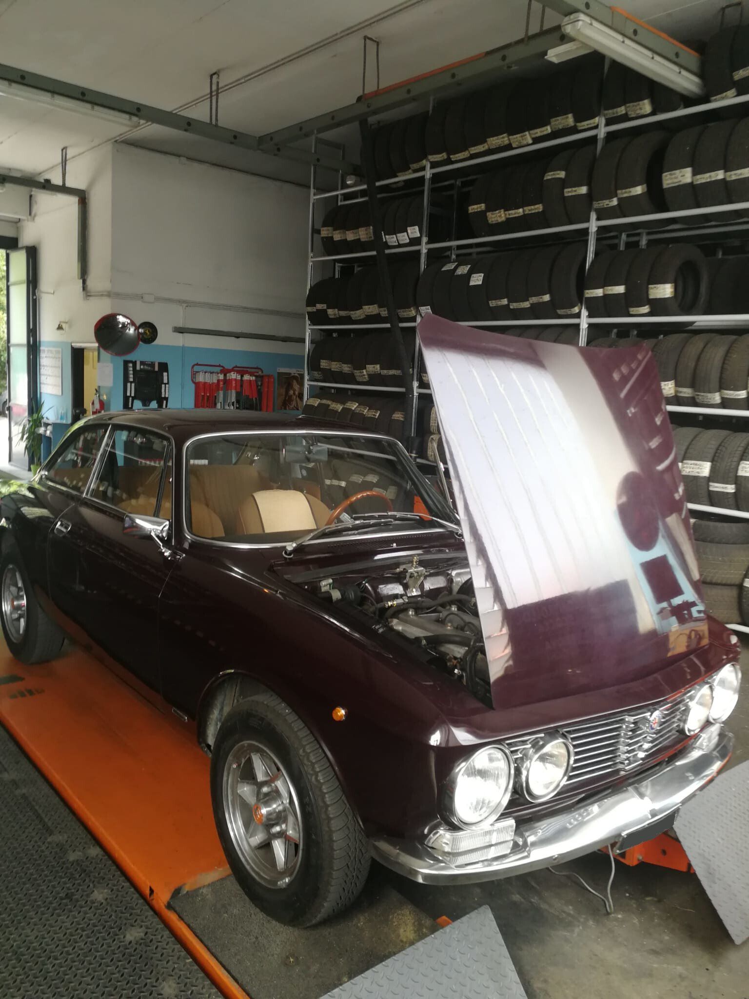 Un'auto d'epoca color bordeaux con il cofano alzato in un garage, accanto a un porta-pneumatici.