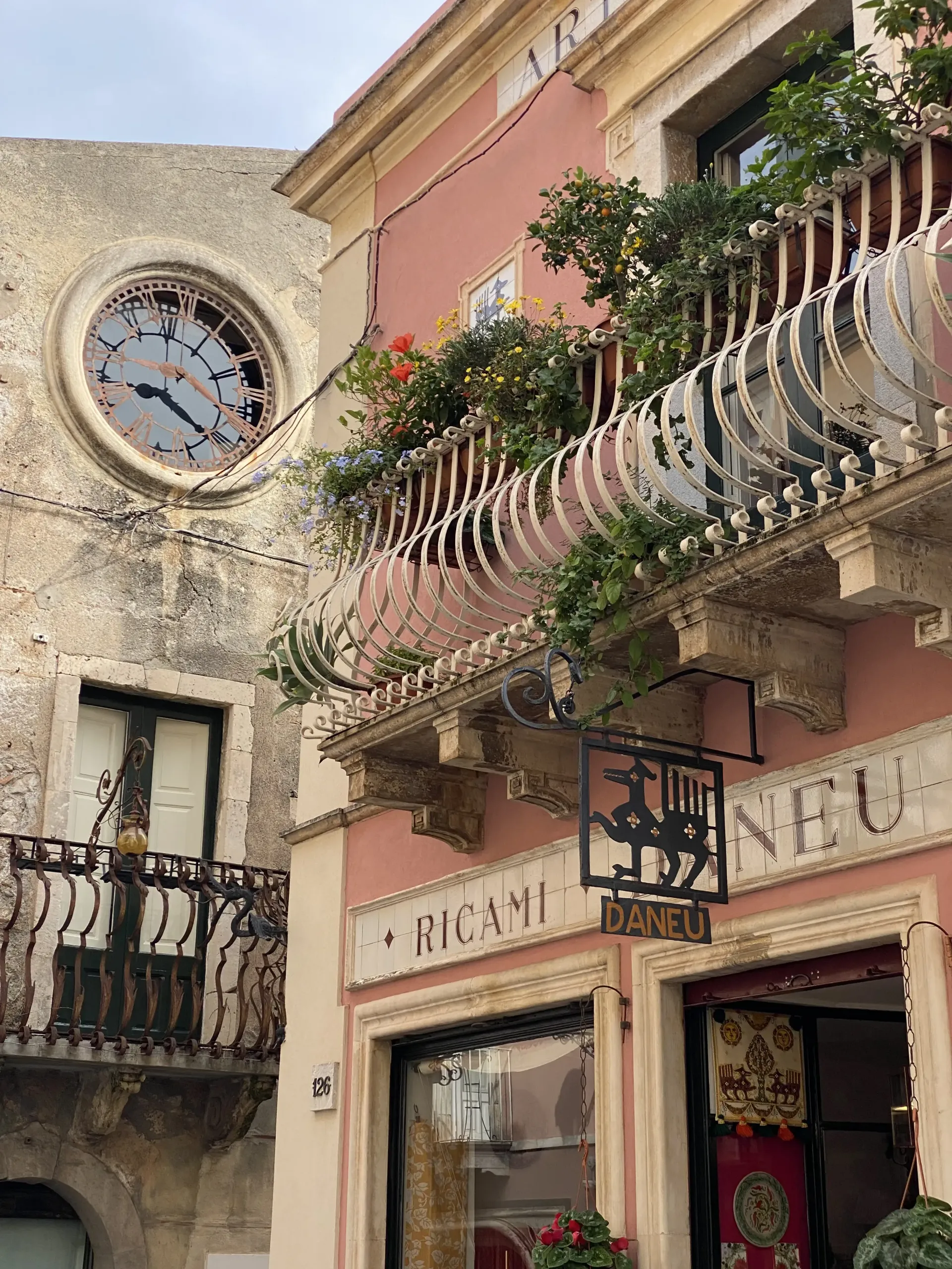 Stretta via con edifici rosa e in pietra, balconi in ferro battuto, un orologio rotondo e un'insegna di un negozio.