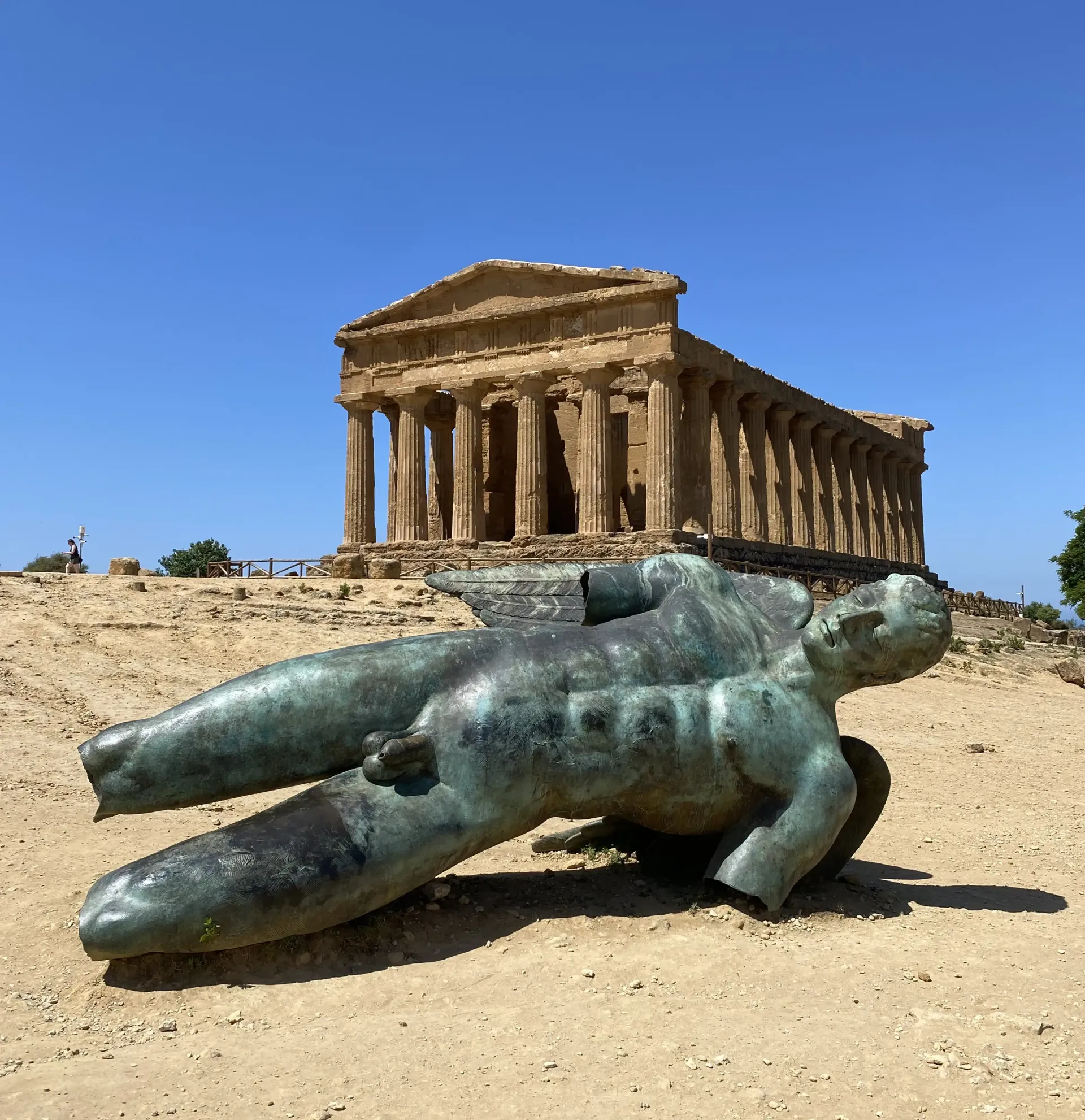 Una gigantesca scultura in bronzo raffigurante una mano davanti a un antico tempio greco, sotto un cielo azzurro e limpido.