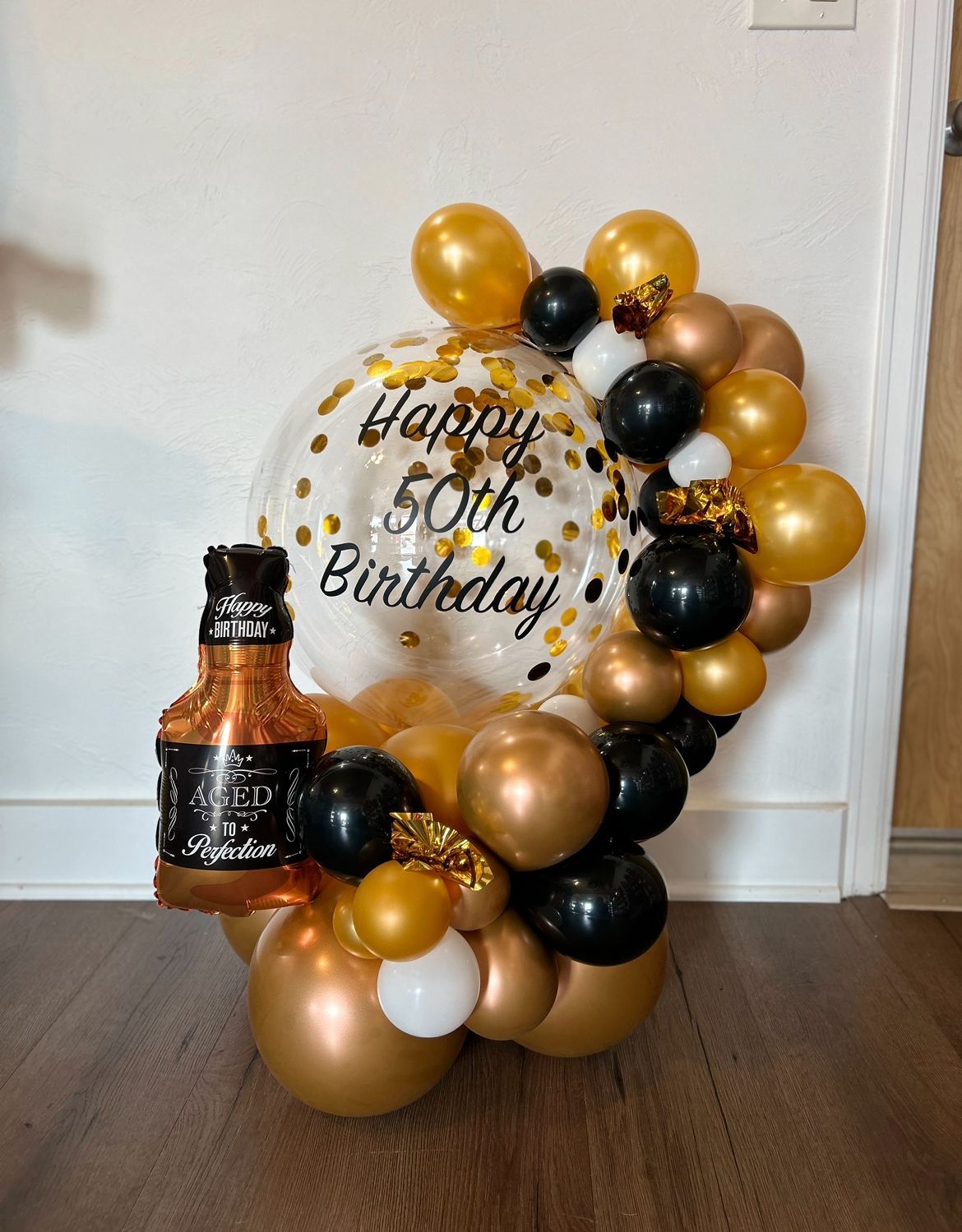 black, gold, and white balloon arrangement for a 50th birthday, featuring a transparent confetti balloon with 'Happy 50th Birthday' text and a foil champagne bottle balloon accent.