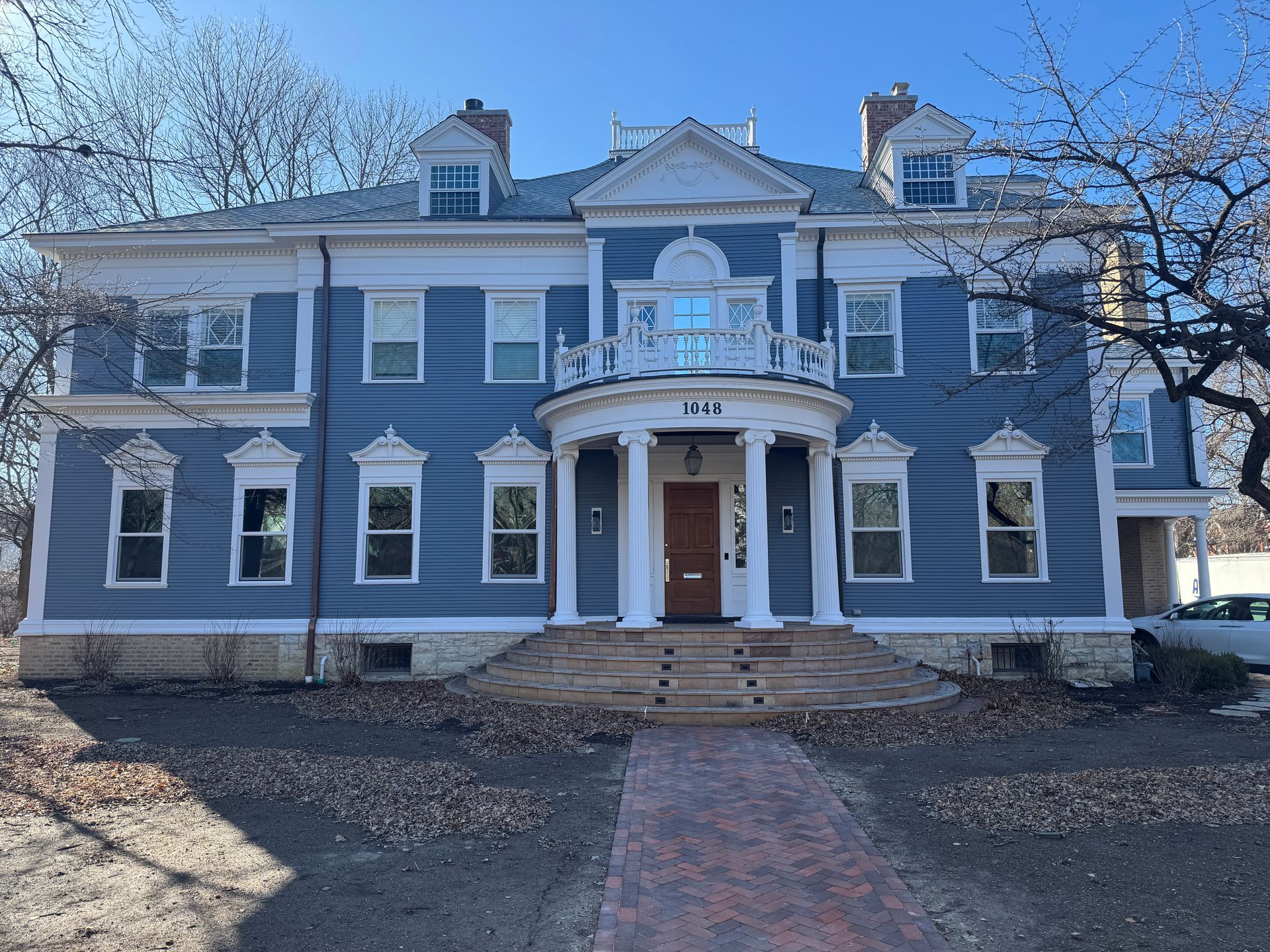 Blue two-story house with white trim and a front porch, set on a brick path with bare trees in the background.