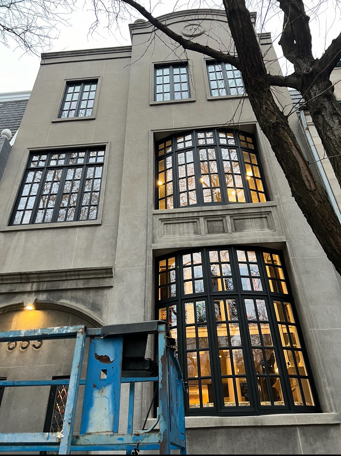 Gray building with black-framed windows. A blue lift sits in front. A tree is to the right.