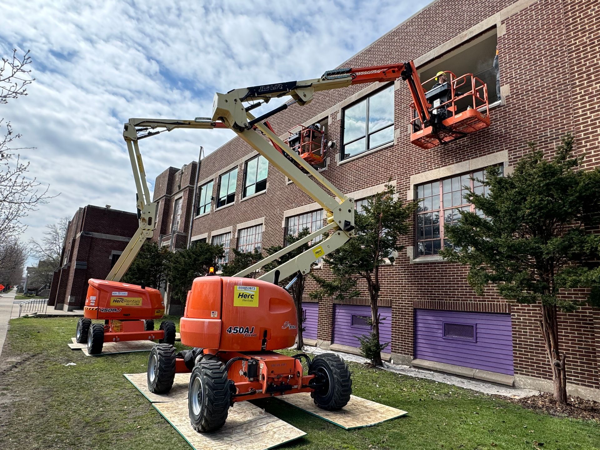Two orange boom lifts by a brick building; workers are replacing windows.