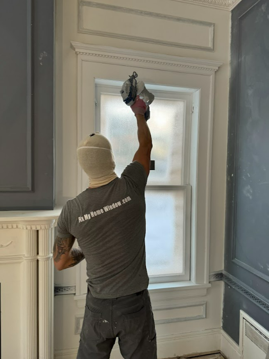 A man in a gray shirt is standing in front of a window.