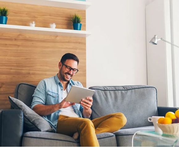 Man sitting on a gray couch using a tablet in a bright living room.