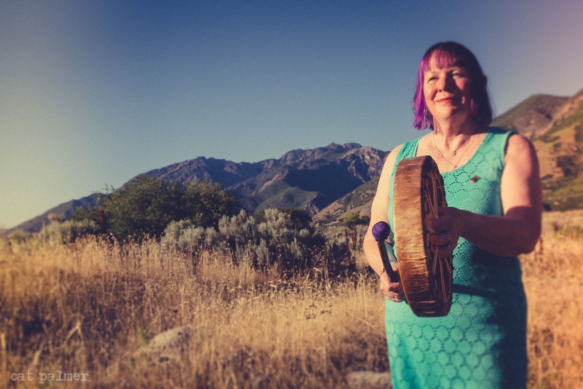Woman with purple hair in turquoise dress, holding a drum in a field with mountains in the background.