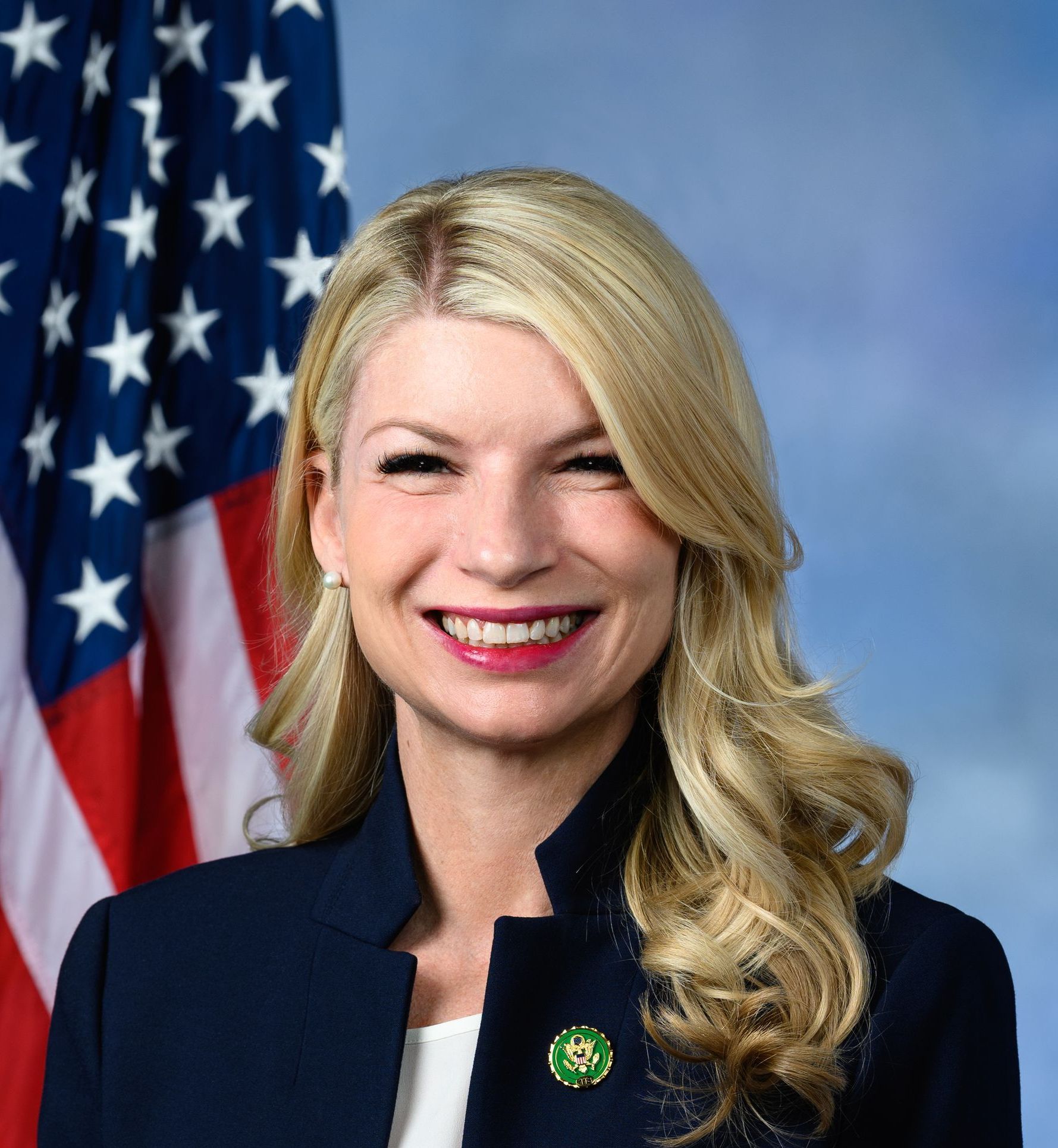 Congresswoman Brittany Pettersen  smiling, wearing a blue blazer, in front of an American flag