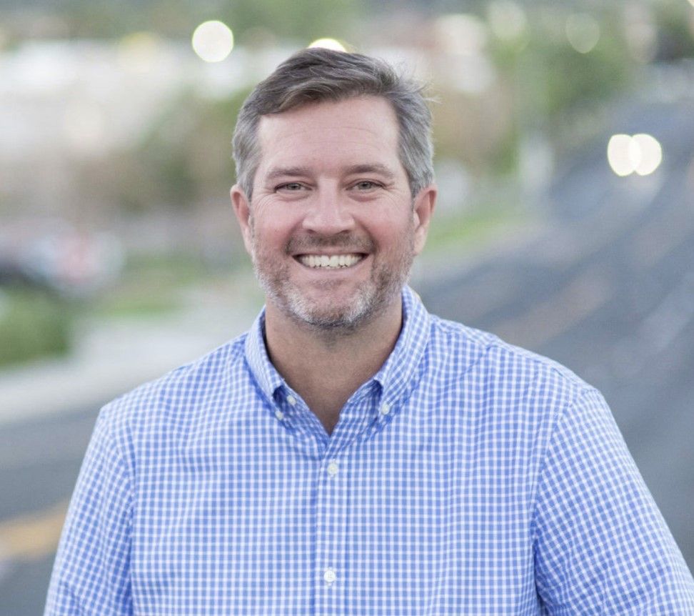 Smiling man in blue checkered shirt; outdoor setting with road and lights.