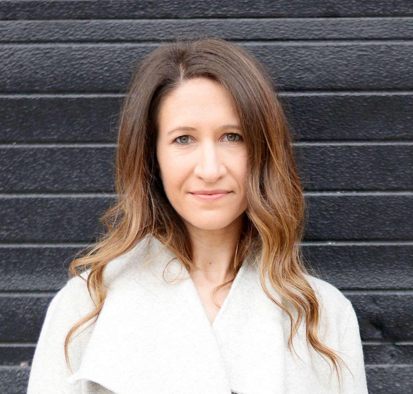 Woman with brown hair, wearing a white coat, smiles in front of a black wall.