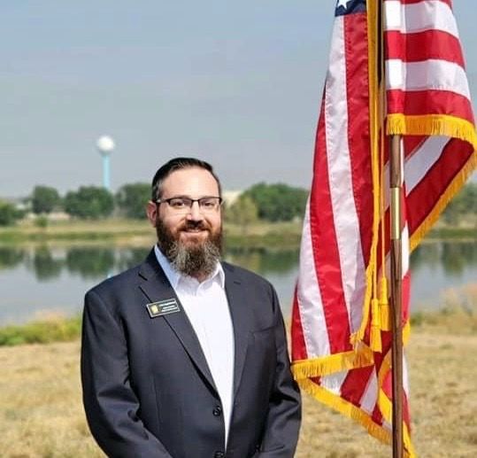 Man in suit with a beard stands by an American flag near water.