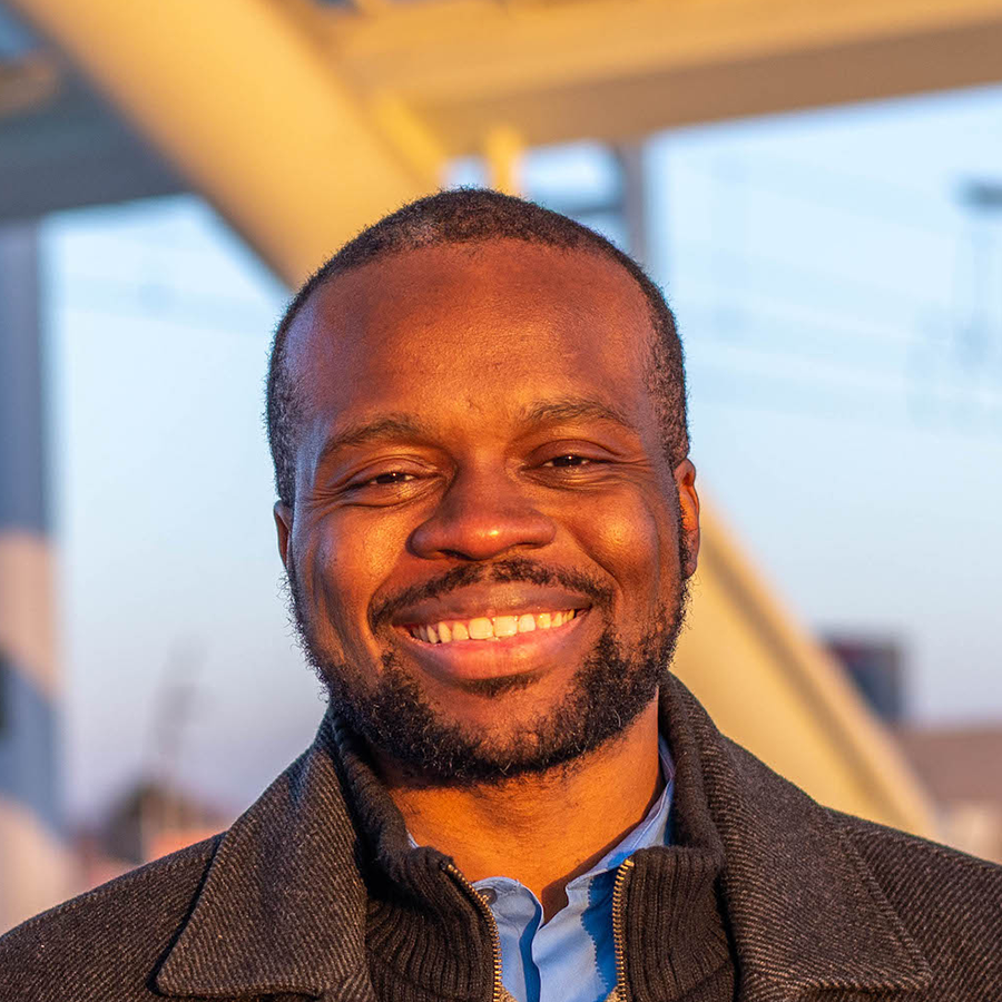Smiling Black man with a short beard, wearing a blue shirt and grey coat, stands outdoors.