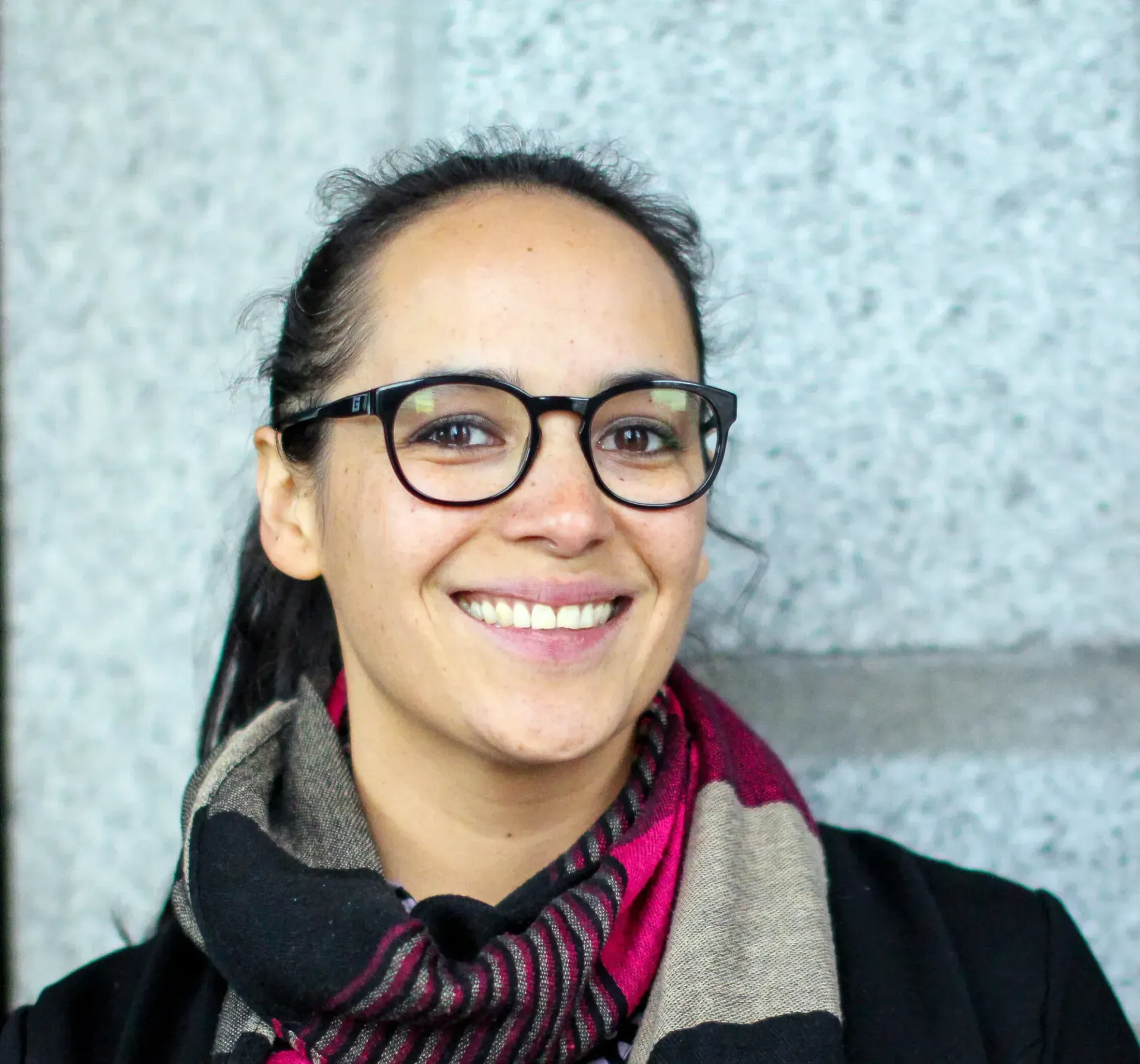 Woman with dark hair, glasses, and scarf, smiling. Leaning against a grey wall.