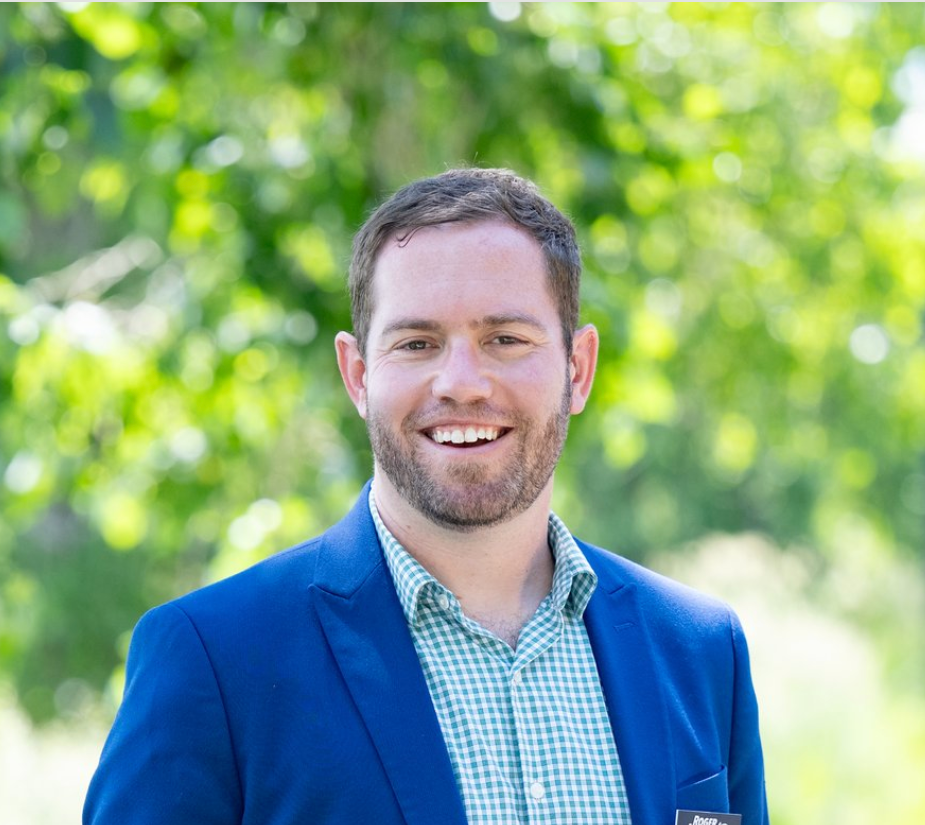 Man in blue blazer and green shirt smiles outdoors.
