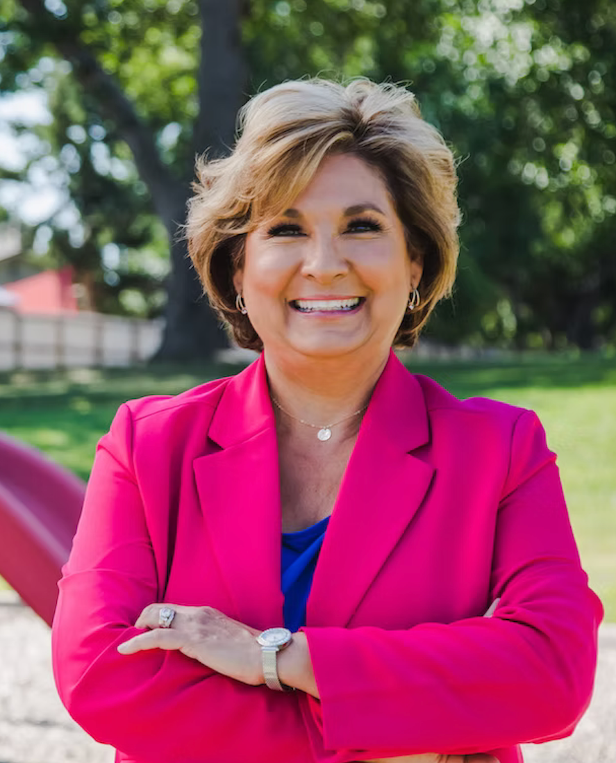 Woman in pink blazer smiles, arms crossed, outdoors. Playground in background.