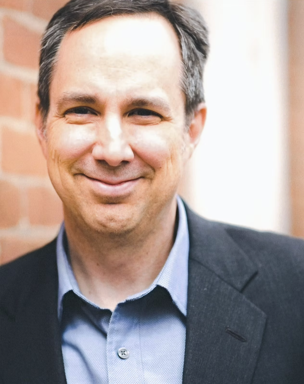 Smiling man in a blue shirt and black blazer, standing near a brick wall.