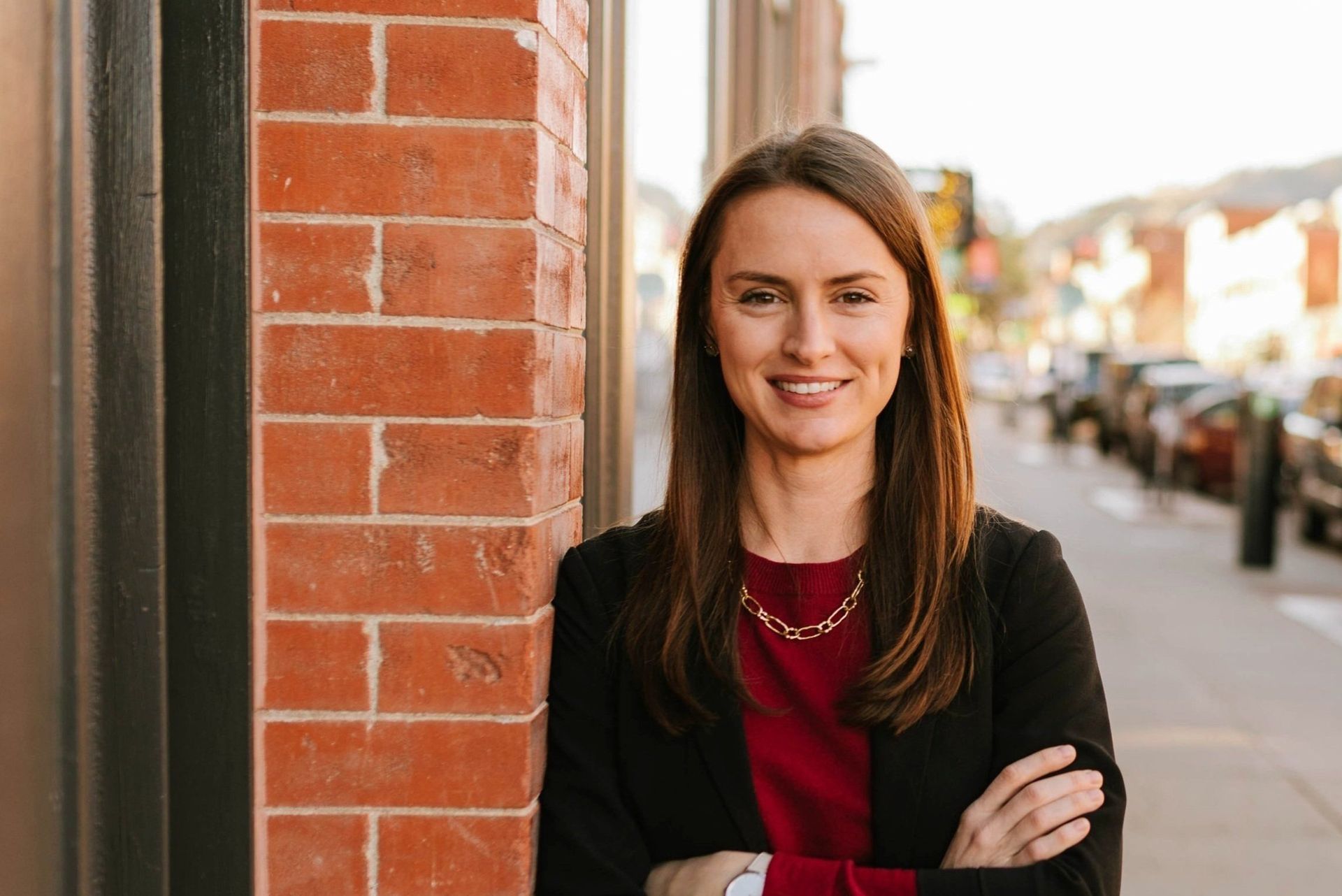 Woman with brown hair in a black blazer and red top, smiling with arms crossed next to a brick wall.