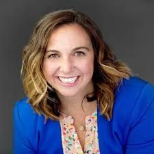 Woman with brown hair, smiling, wearing a blue blazer and floral print shirt.