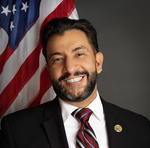 Man with a beard, smiling in a suit, in front of the American flag.