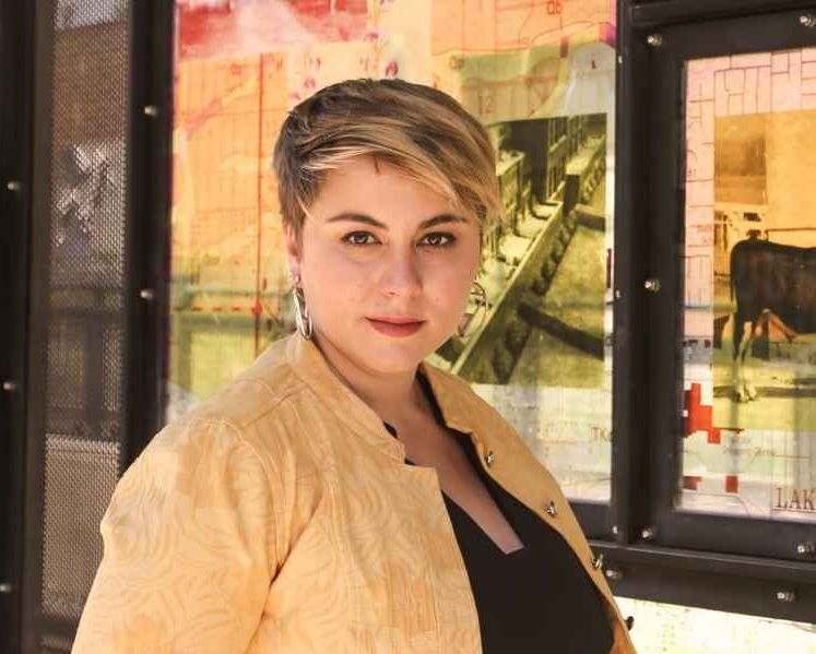 Woman with short blonde hair in front of an art piece, wearing a yellow jacket and black top.