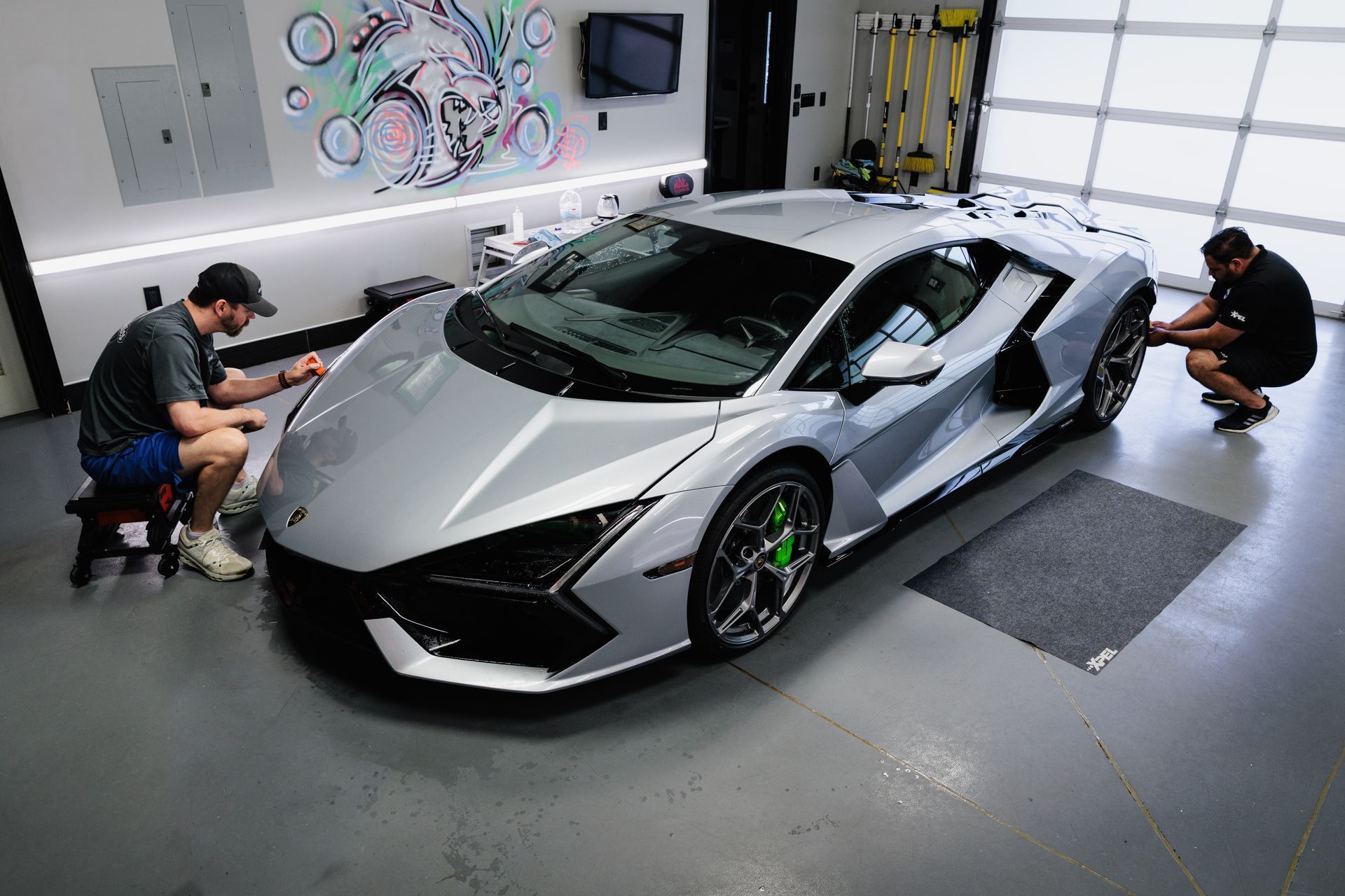 Two detailers polishing a silver sports car in a garage-like showroom