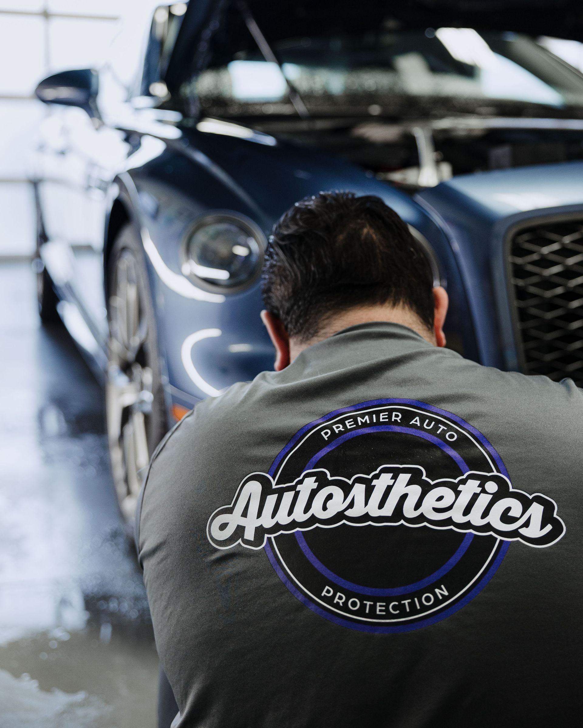 Detailer polishing a blue car beside an Autosthetics logo on a gray shirt