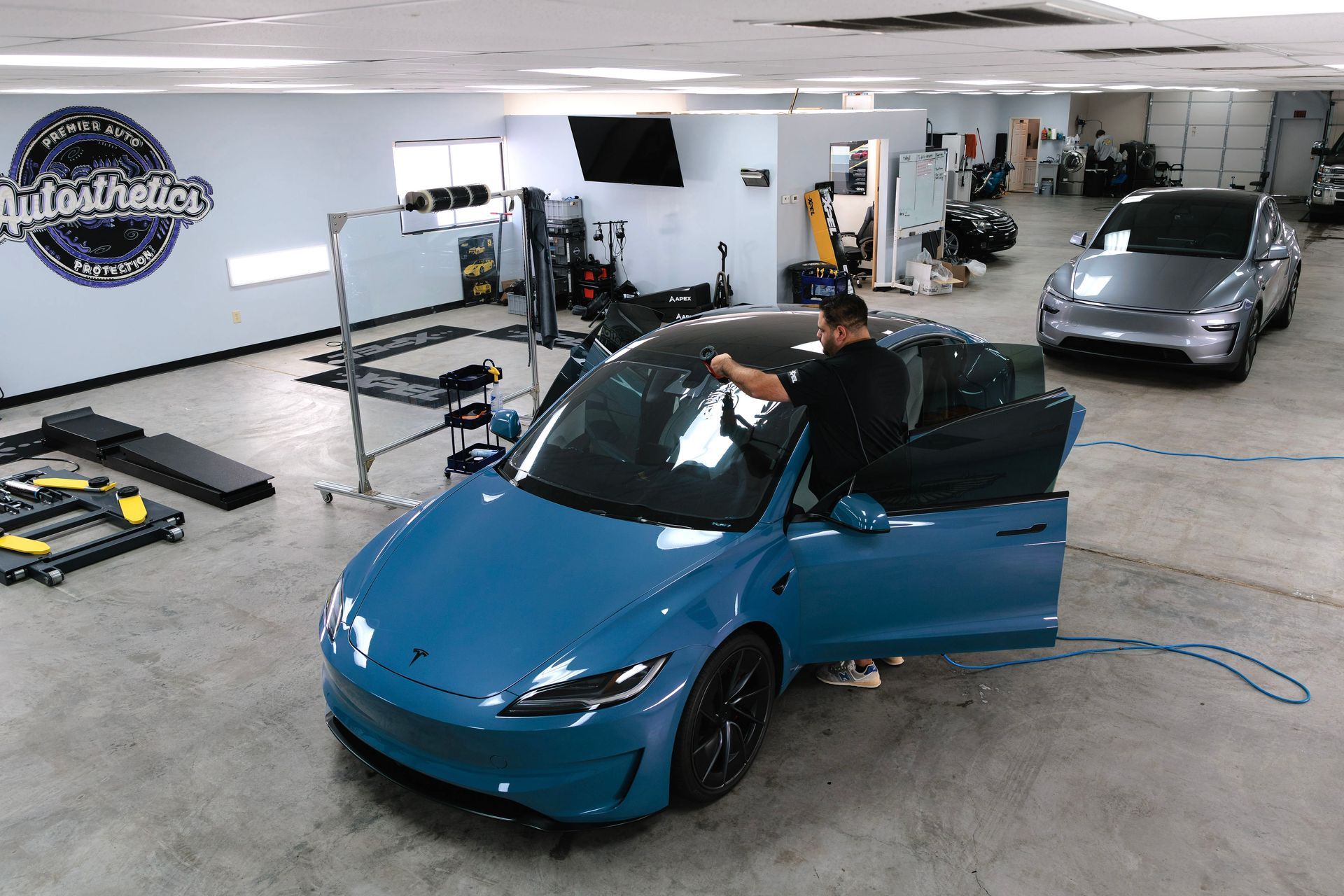 Blue sports car being worked on in an auto shop, with a gray car parked in the background.