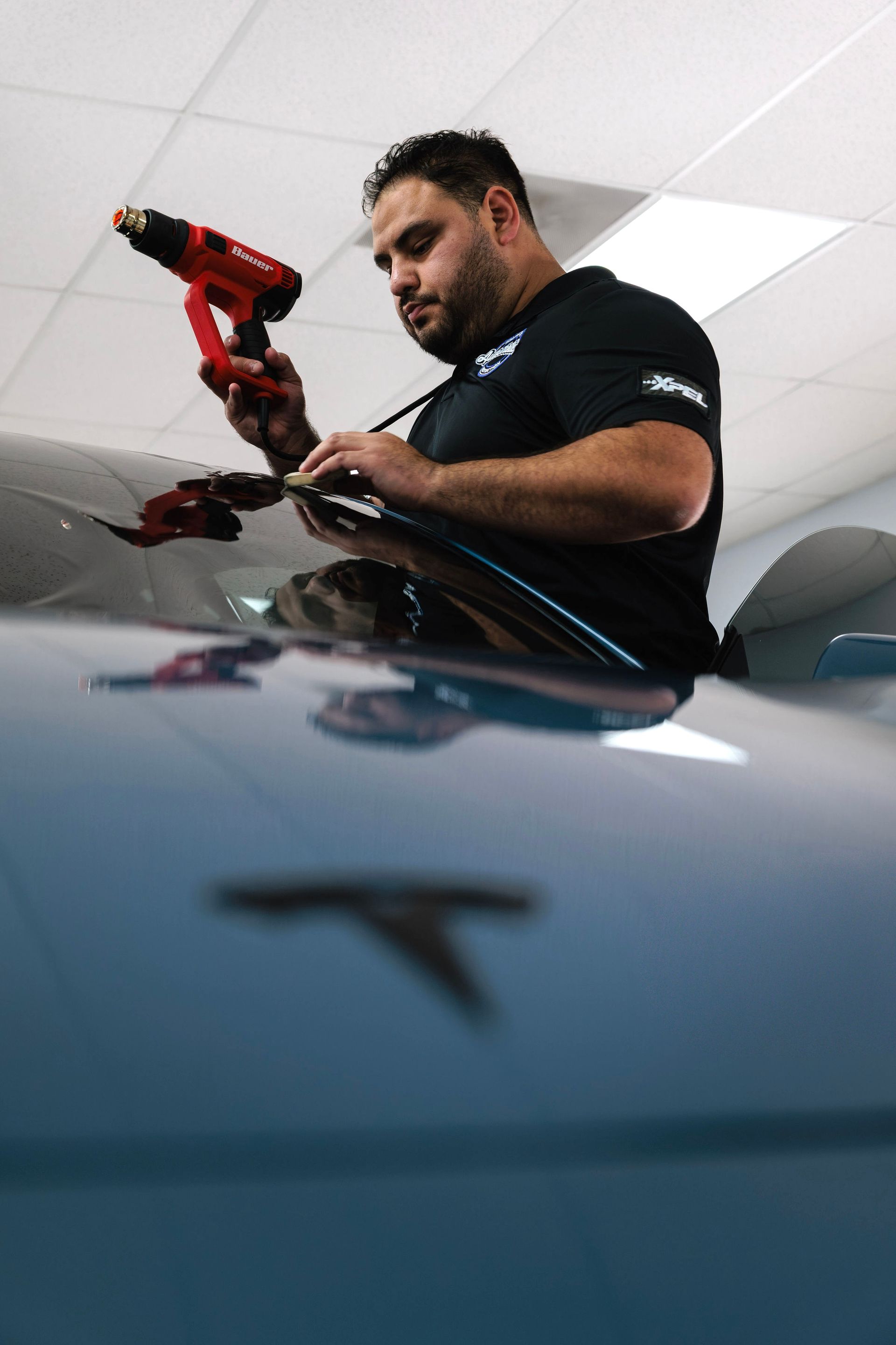 Man using a red drill to work on a dark car in a garage, viewed from a low angle
