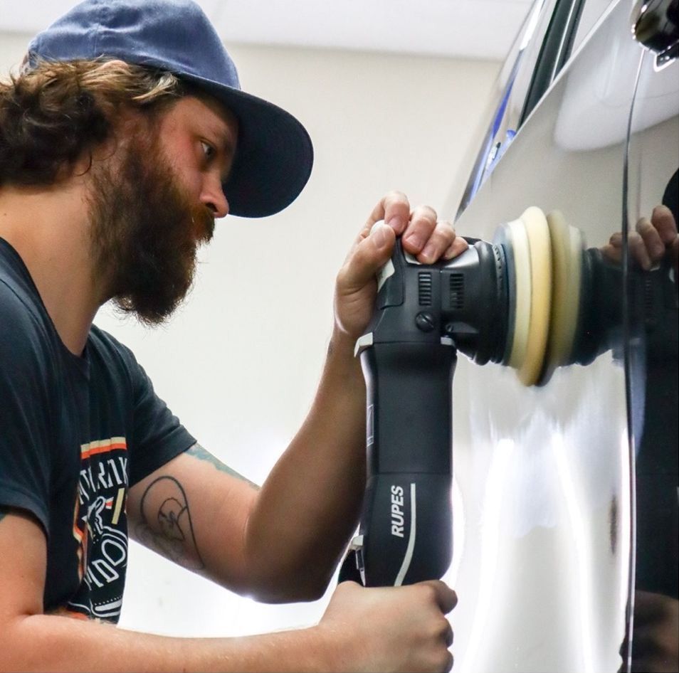 Man using a polishing machine on a black car. He wears a hat and focuses on the work in a bright setting.