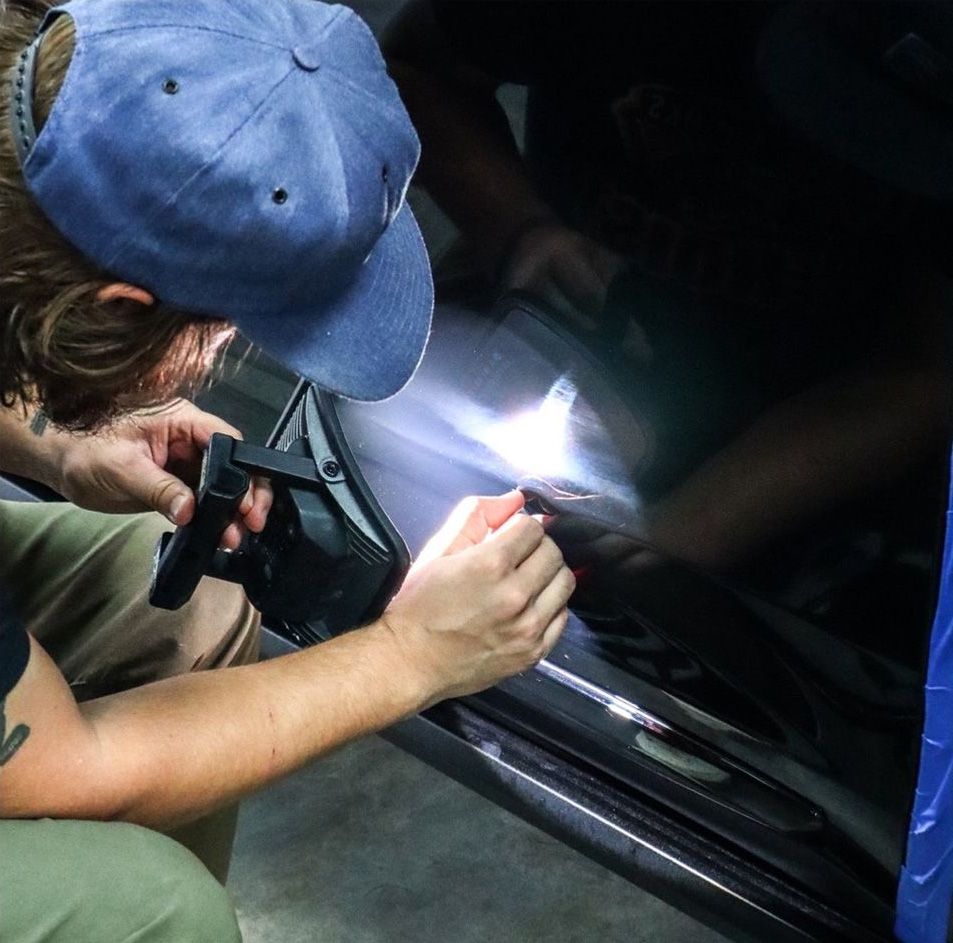 Person sprays liquid on a black car door, inspecting the surface with a bright light.