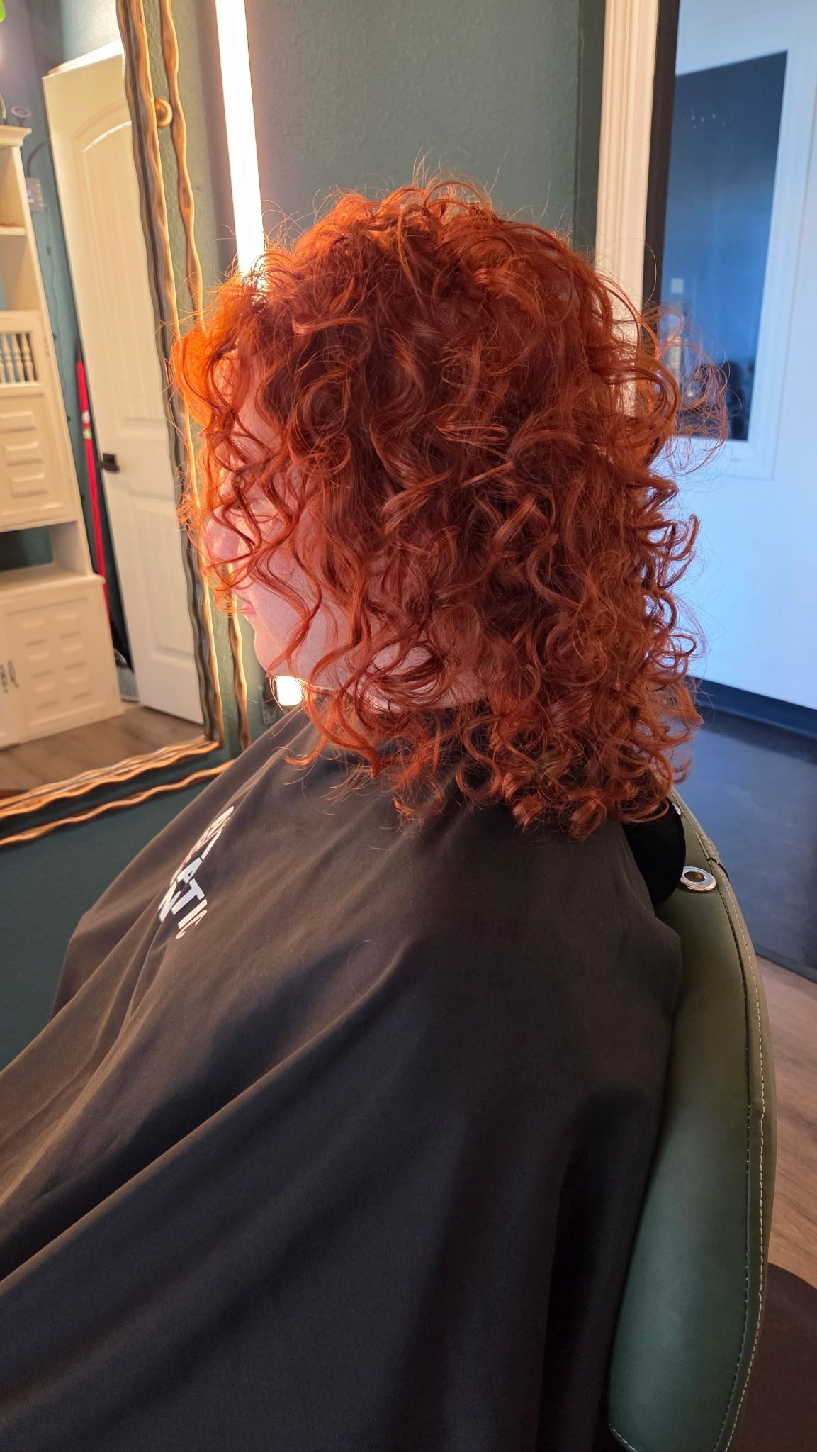 A person with vibrant ginger, shoulder-length curly hair sits in a salon chair wearing a black cape.