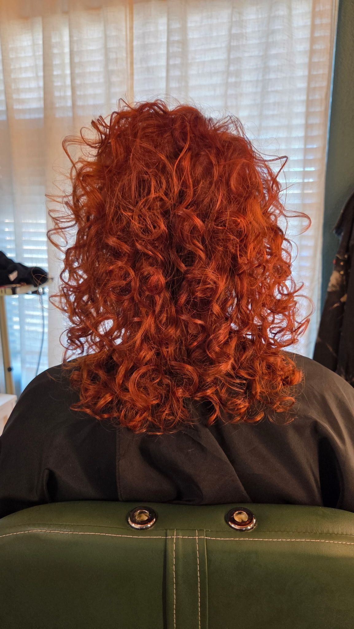 A person with shoulder-length, vibrant red curly hair, sitting in a salon chair in front of a white window curtain.