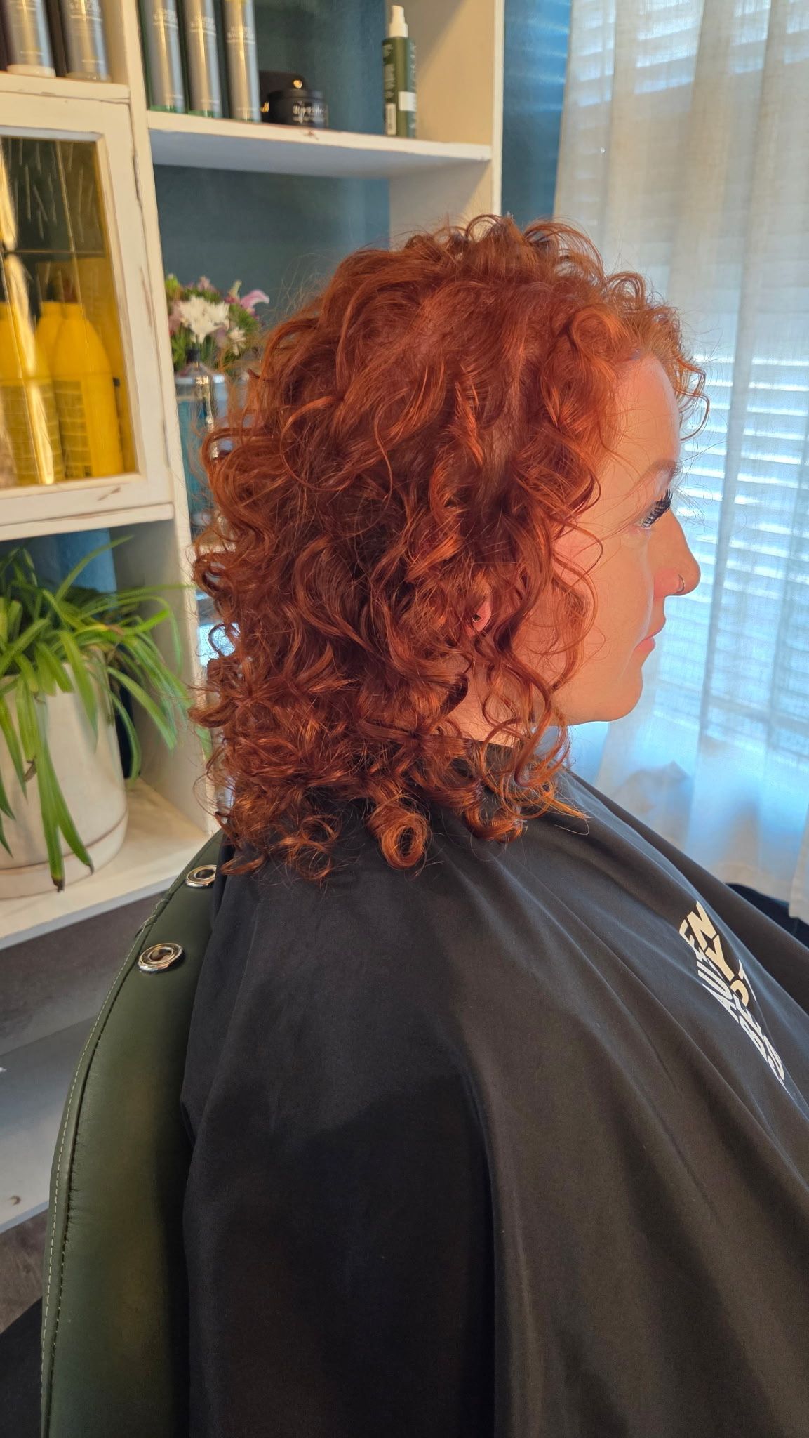 A person with shoulder-length, vibrant copper curly hair sits in a salon chair wearing a black cape.