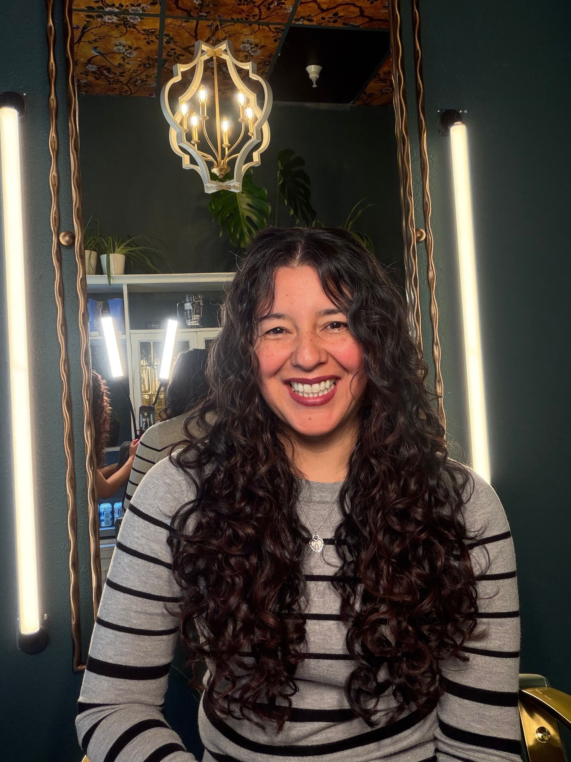 A smiling person with long, dark, curly hair wearing a gray and black striped long-sleeve shirt in a salon setting.