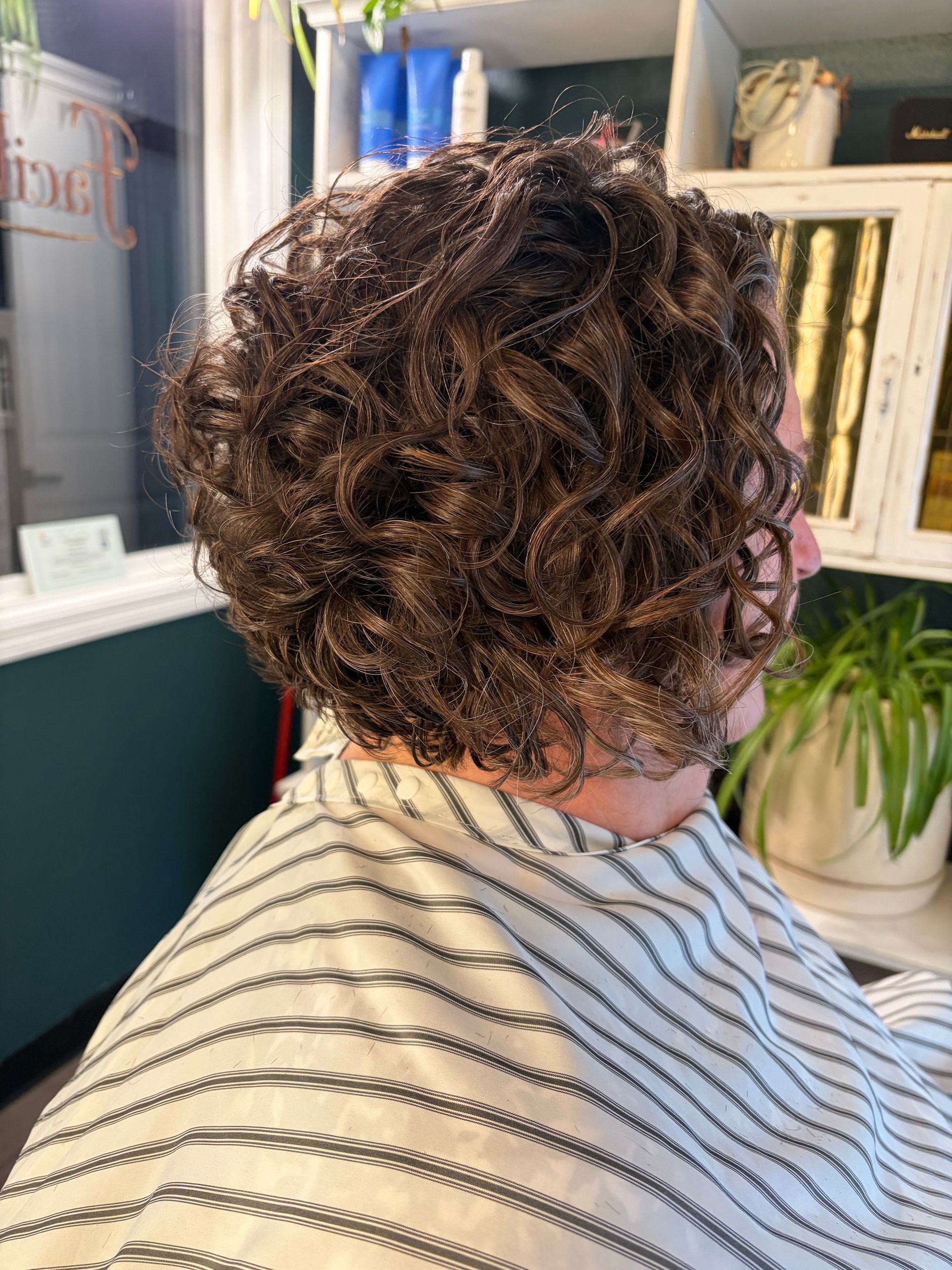 A person with dark, curly hair shown from the side in a salon setting, wearing a striped cape.