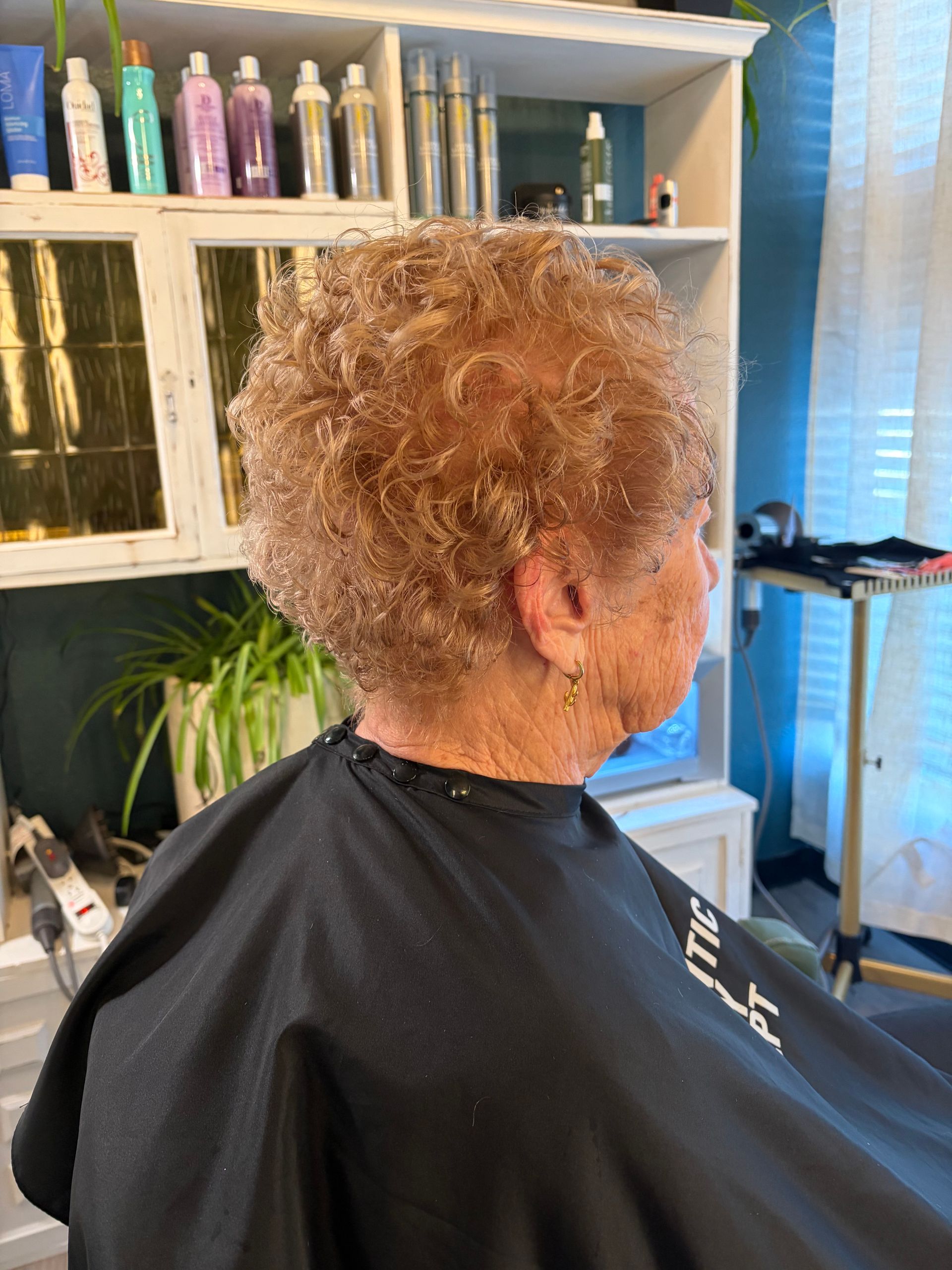 A person with short, curly, light-brown hair sits in a salon chair wearing a black cape, with shelves of products behind.