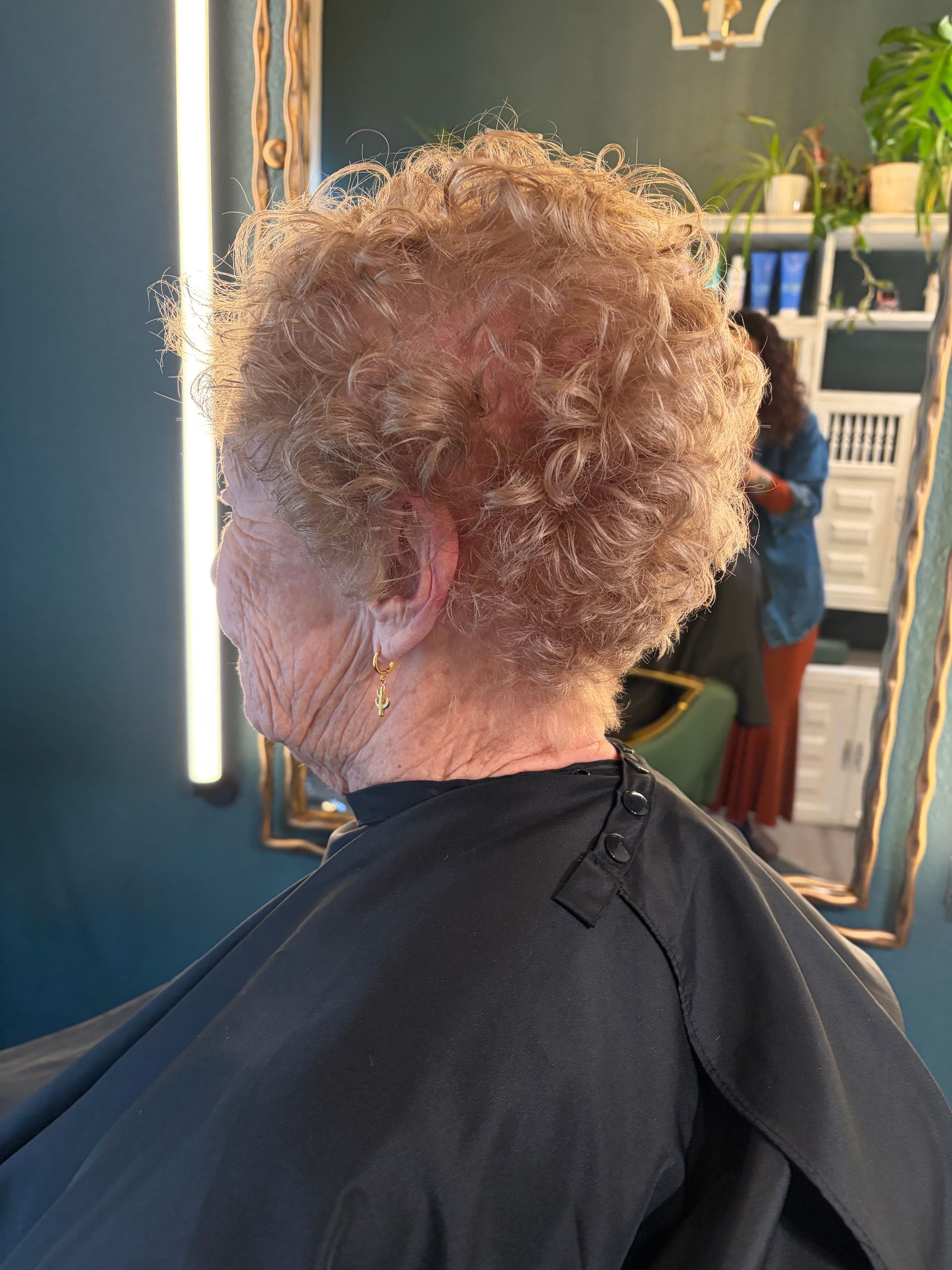 A person with short, curly, light brown hair sits in a salon chair wearing a black cape, seen from a side profile.