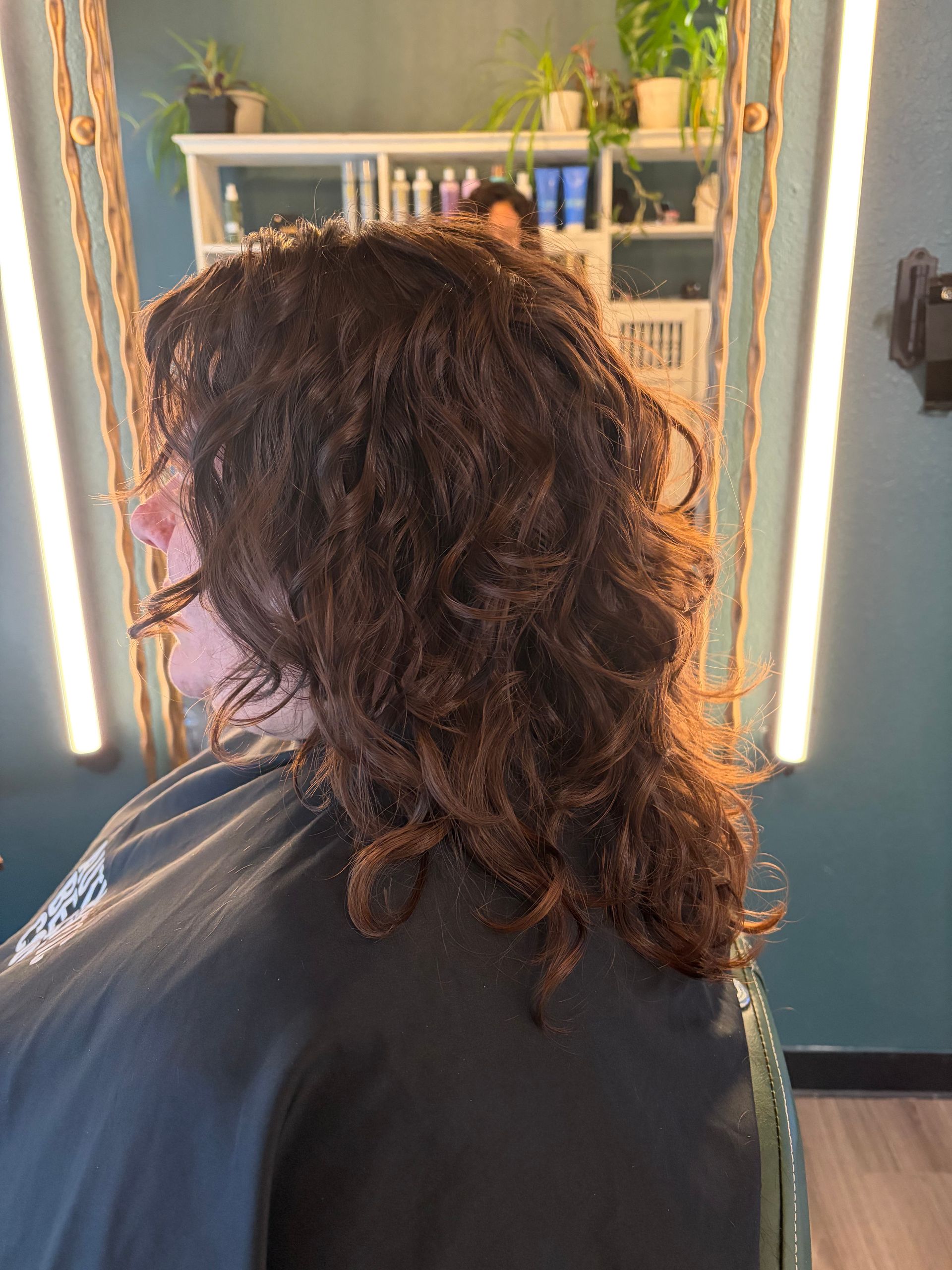A person with long, dark, wavy hair sitting in a salon chair in front of a mirror with white shelving and plants.