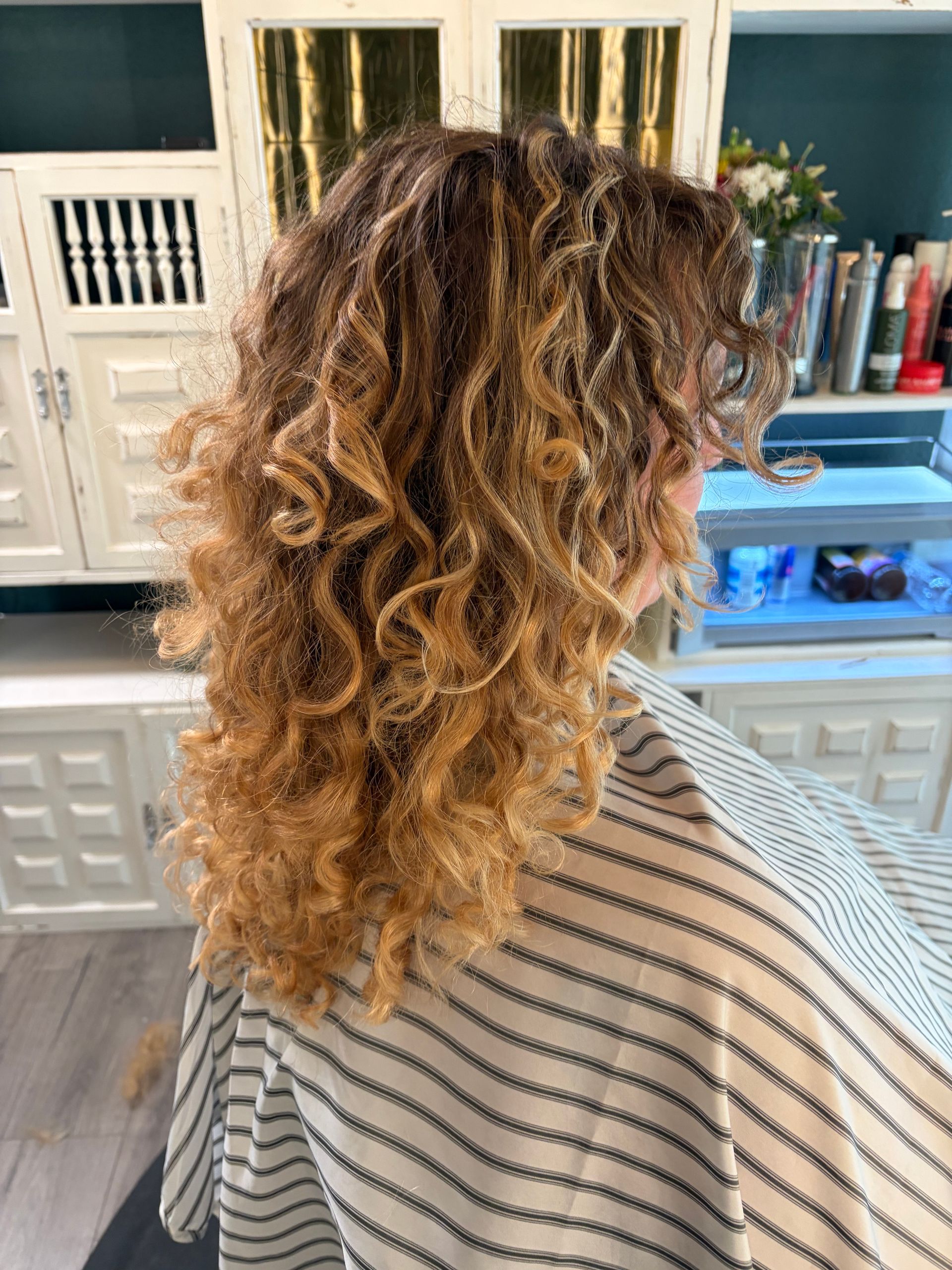 A person with long, curly, highlighted brown and blonde hair wearing a striped salon cape in a hair salon.