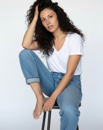 A person with dark curly hair, wearing a white V-neck t-shirt and blue jeans, sits on a stool against a white background.